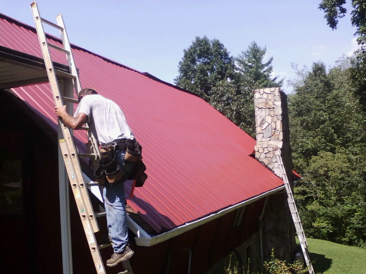 Roofer on a ladder beside a red metal roof and stone chimney — Soffit & Fascia Repair by Roofing Brothers in Agra